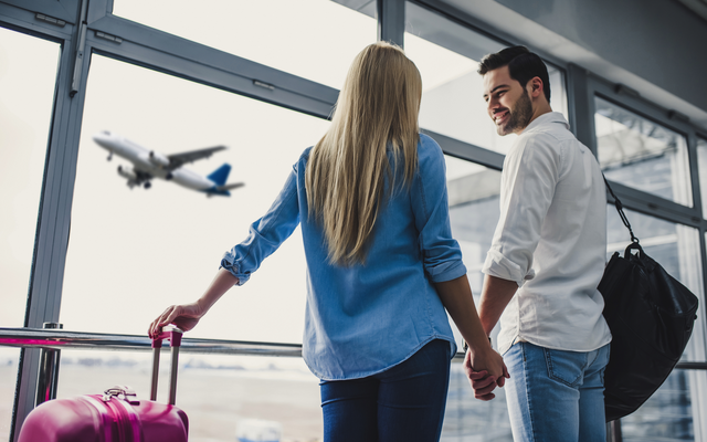 Paar hält sich am Flughafen an den Händen, Flugzeug fliegt im Hintergrund.