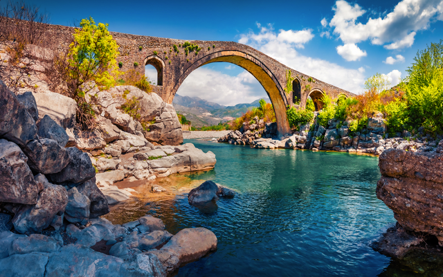 Steinbrücke über einen Fluss in malerischer Landschaft bei sonnigem Wetter.