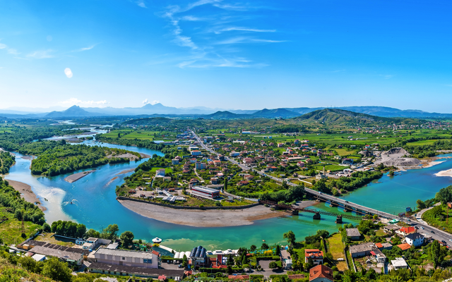 Panorama einer Flusslandschaft mit Wiesen, Bergen und einer Kleinstadt.