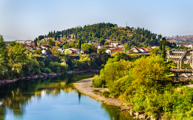 Landschaft mit Fluss, bewachsenem Hügel und Dorf im Hintergrund.