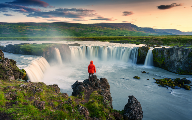 Person in roter Jacke steht vor einem Wasserfall in einer grünen Landschaft bei Sonnenuntergang.