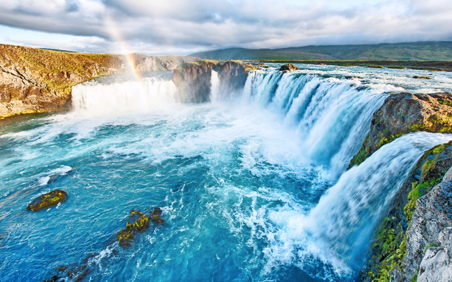 Breiter Wasserfall mit starkem Wasserfluss, umgeben von grüner Landschaft und Regenbogen.