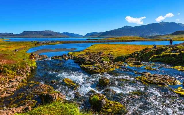 Landschaft in Island mit Fluss und Bergen im Hintergrund, bei sonnigem Wetter und blauem Himmel.