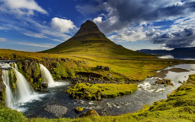 Berglandschaft mit Wasserfall und grünem Tal unter bewölktem Himmel.