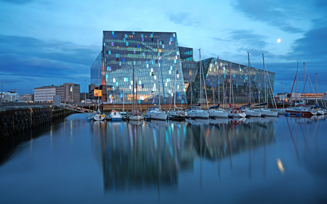 Modernes Glasgebäude am Wasser mit Segelbooten im Vordergrund.