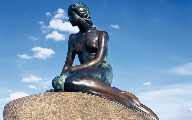 Statue der kleinen Meerjungfrau auf einem Stein sitzend mit blauem Himmel im Hintergrund.
