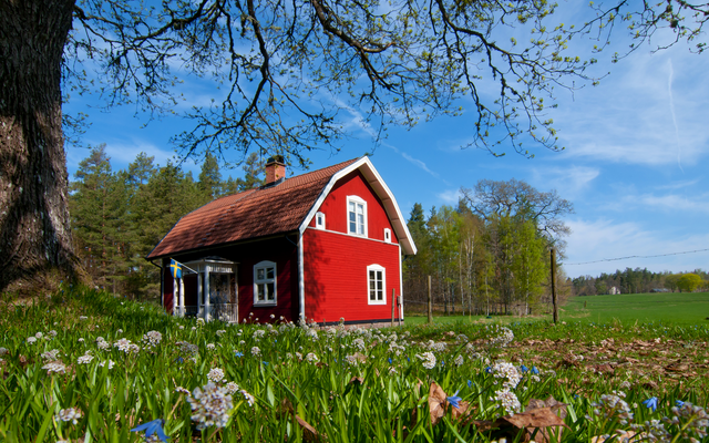 Rotes Landhaus auf Wiese, umgeben von Bäumen und blühenden Blumen.