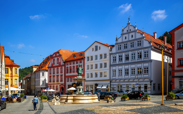 Historischer Marktplatz mit farbigen Gebäuden und Brunnen bei blauem Himmel.
