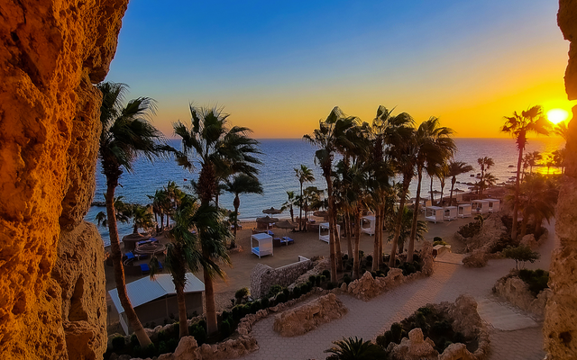 Blick aus einem Felsenfenster auf tropischen Strand bei Sonnenuntergang mit Palmen.