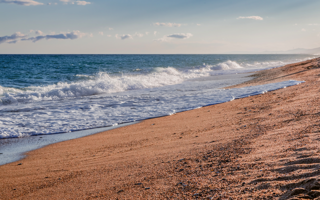Wellen am sandigen Strand bei blauem Himmel und leicht bewölktem Horizont.