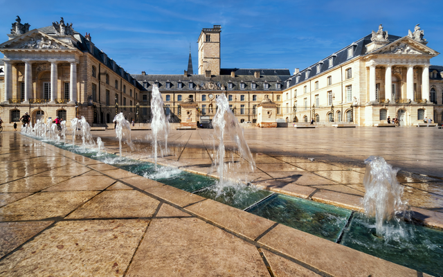Platz mit Brunnen vor historischem Gebäude bei sonnigem Wetter.