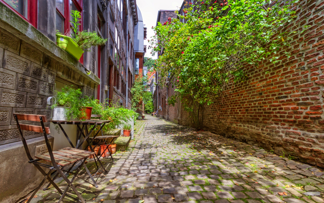 Schmale gepflasterte Gasse mit Tisch und Stühlen, umgeben von Backsteinmauern und Pflanzen.