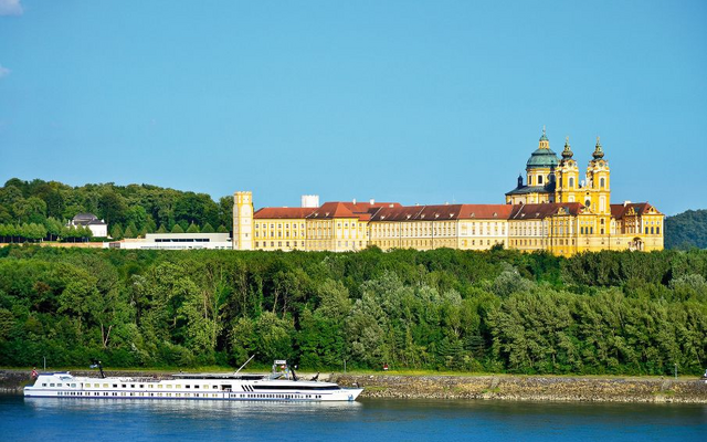 Barockes Kloster auf Hügel über Fluss, mit vorbeifahrendem Kreuzfahrtschiff.
