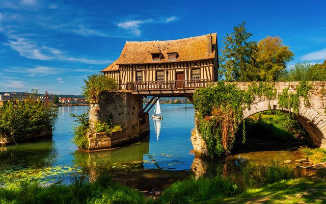 Fachwerkhaus auf einer alten Steinbrücke über einem Fluss mit Segelboot im Hintergrund.