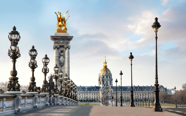 Brücke mit Laternen und goldener Statue vor Gebäude mit Kuppel im Hintergrund.