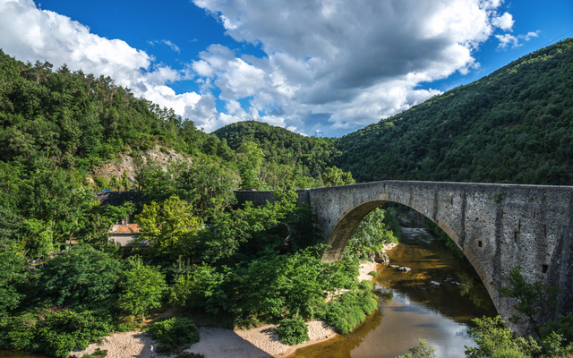 Steinbrücke über Fluss in bewaldeter Hügellandschaft bei blauem Himmel.